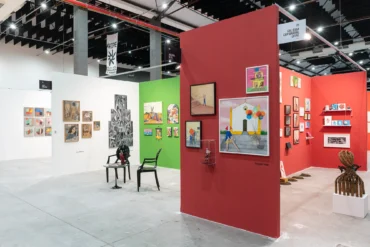Estandes de galerias em feira de arte, com paredes coloridas em branco, verde e vermelho exibindo quadros, esculturas e objetos diversos sob estrutura industrial de teto. Fotos Danilo Galvão