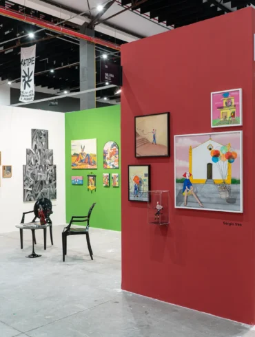 Estandes de galerias em feira de arte, com paredes coloridas em branco, verde e vermelho exibindo quadros, esculturas e objetos diversos sob estrutura industrial de teto. Fotos Danilo Galvão