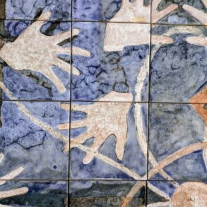 Painel de azulejos artesanais com fundo azul mosqueado e mãos estilizadas em tons terracota, bege e marrom sobrepostas, formando composição abstrata.