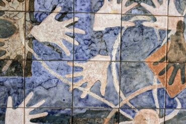 Painel de azulejos artesanais com fundo azul mosqueado e mãos estilizadas em tons terracota, bege e marrom sobrepostas, formando composição abstrata.