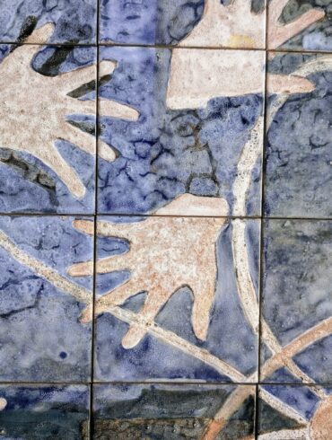 Painel de azulejos artesanais com fundo azul mosqueado e mãos estilizadas em tons terracota, bege e marrom sobrepostas, formando composição abstrata.