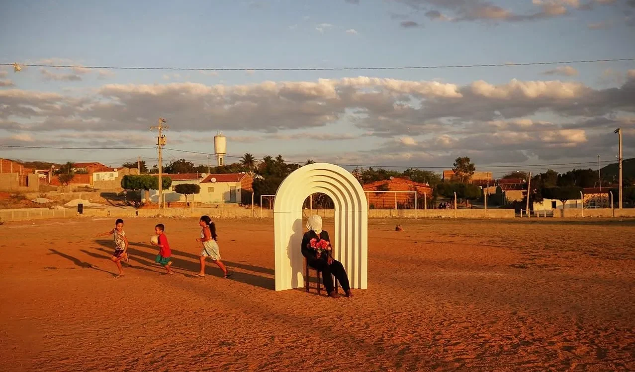 Em um campo de terra batida ao entardecer, uma pessoa sentada com flores no colo sob um arco branco escultural, enquanto crianças correm ao lado com uma bola.