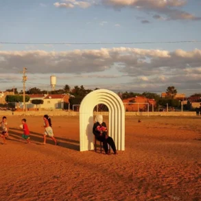 Em um campo de terra batida ao entardecer, uma pessoa sentada com flores no colo sob um arco branco escultural, enquanto crianças correm ao lado com uma bola.
