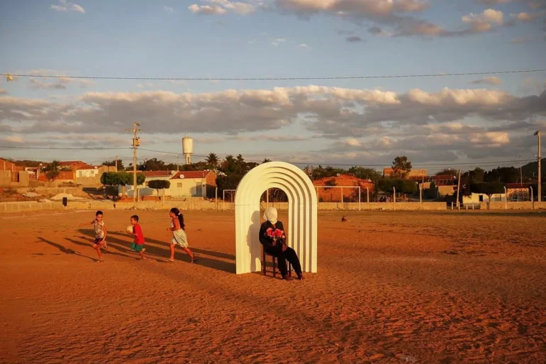 Em um campo de terra batida ao entardecer, uma pessoa sentada com flores no colo sob um arco branco escultural, enquanto crianças correm ao lado com uma bola.